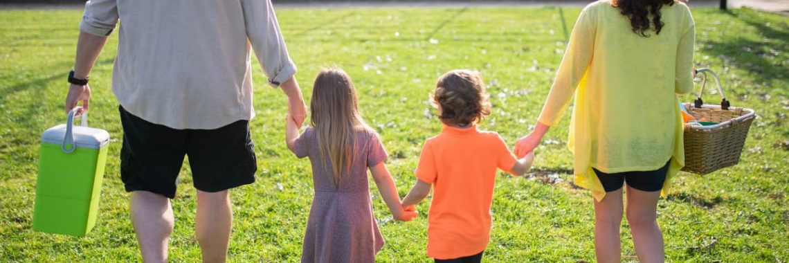 A family of four walking hand in hand through a sunny park carrying picnic supplies.