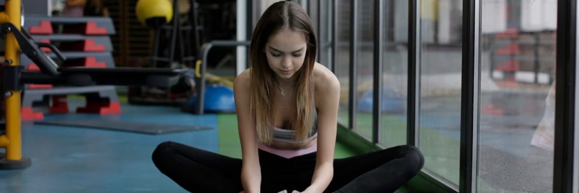 A woman performing a stretching routine inside a gym next to large windows.