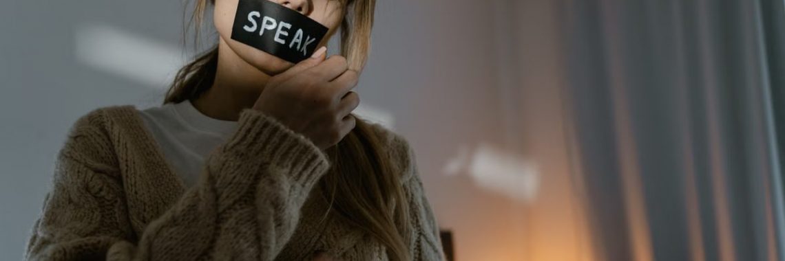 A woman standing in a dimly lit room removing tape with 'SPEAK' written, symbolizing empowerment.